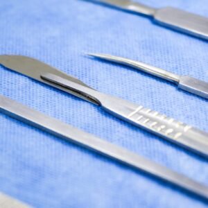 Instrumental quirúrgico de acero inoxidable sobre un paño azul, incluyendo un bisturí, pinzas y tijeras.