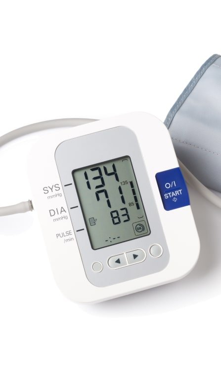 Tensiómetro digital blanco con una pantalla que muestra una lectura de presión arterial de 134/71 y un pulso de 83, junto a su brazalete gris.