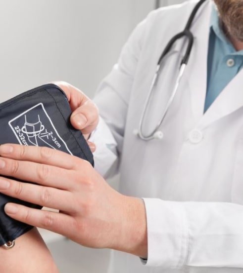 Médico con bata blanca y estetoscopio midiendo la presión arterial de un paciente con un esfigmomanómetro manual negro.