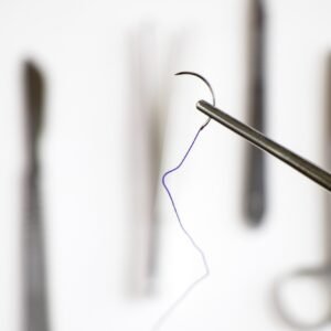 Pinza quirúrgica sosteniendo una aguja curva con hilo de sutura, con instrumentos médicos desenfocados en el fondo.