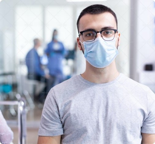 Hombre con gafas y mascarilla azul, vistiendo una camiseta gris, de pie en un entorno médico con personal de salud desenfocado en el fondo.